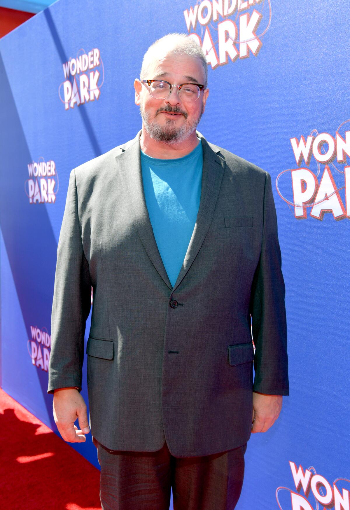 Ken Hudson smiles and poses while wearing a dark gray suit and a blue shirt and glasses