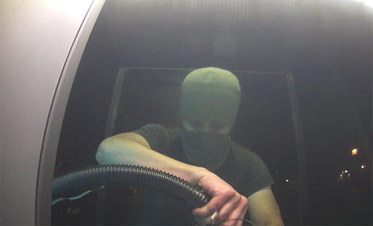 A man is seen behind the wheel of a vehicle, with a mask and cap covering all of his face except for his eyes.