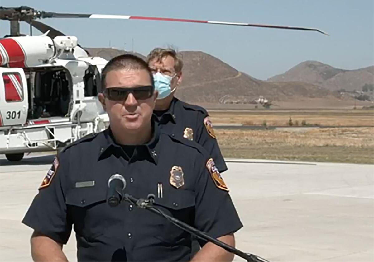 A man in dark uniform, wearing sunglasses, speaks at a microphone with a helicopter in the background.