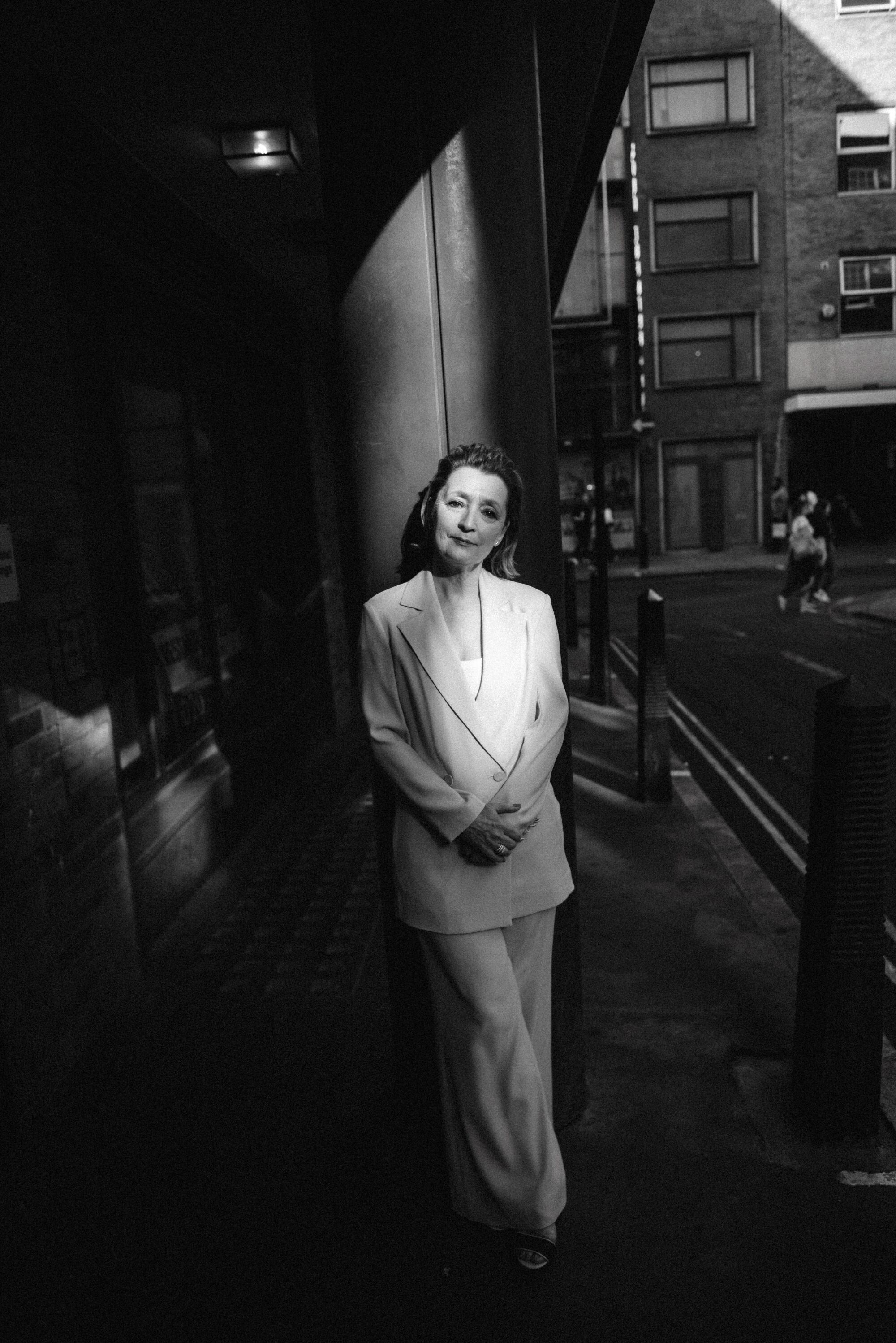 A woman in a white suit leans against a pole outdoors as sunshine hits her