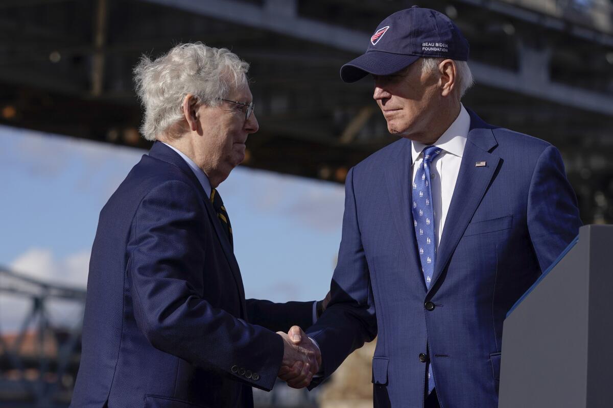 Two white-haired men in blue suits shake hands. One is wearing a baseball cap.