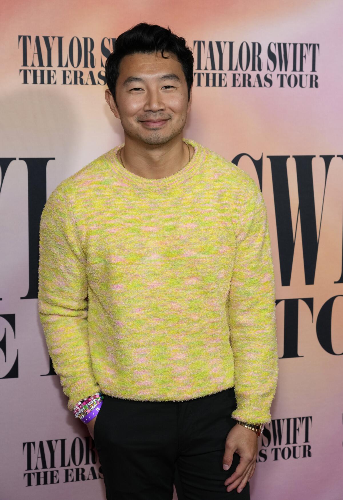 Ο Simu Liu χαμογελά και ποζάρει με ένα κίτρινο πουλόβερ 