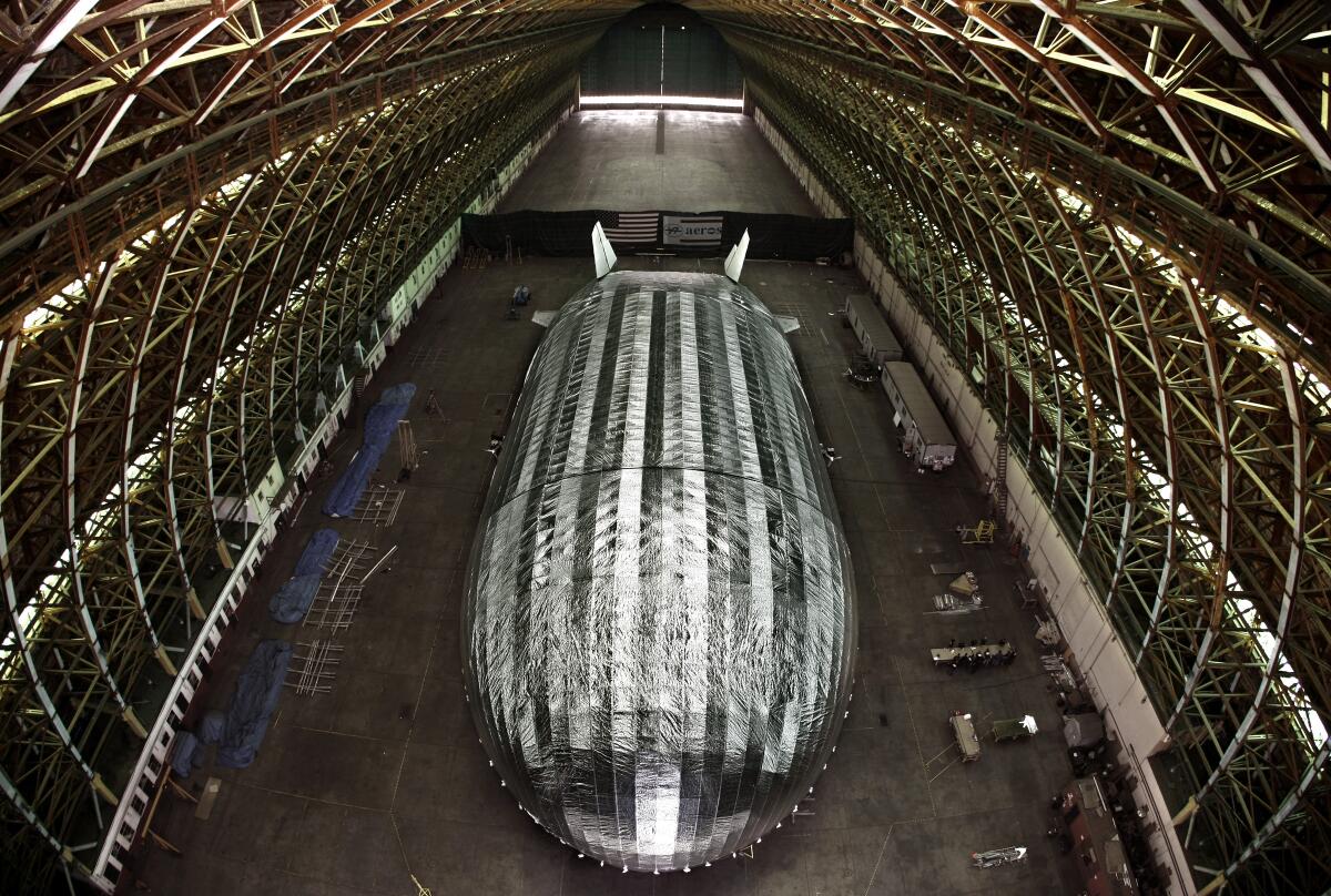 A metallic-looking blimp-like airship under construction inside a cavernous aircraft hangar