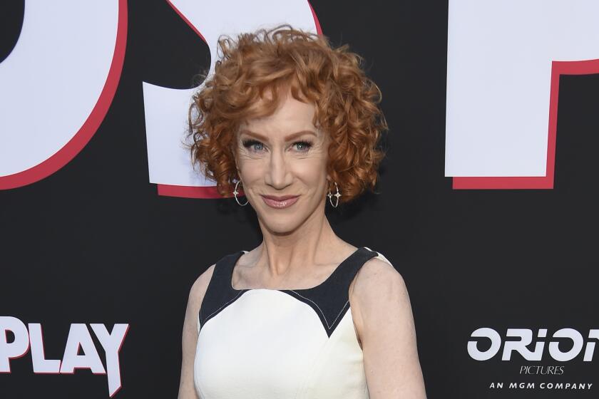 Kathy Griffin smiling with short curled red hair and wearing white and black dress, hoop earings at red carpet event