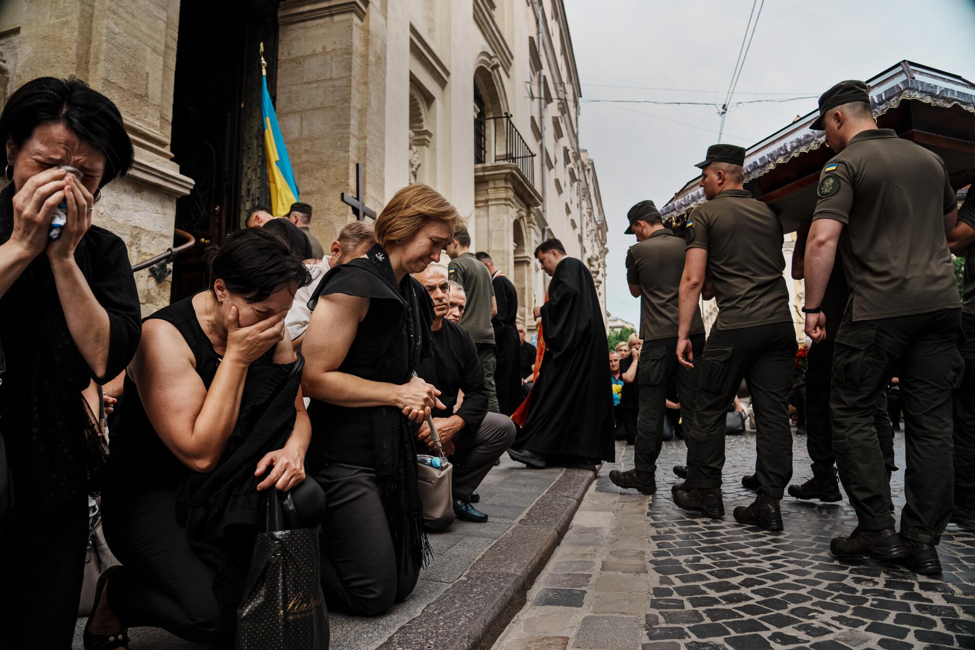 Mourners kneel and weep as a funeral procession, with soldiers carrying a casket, passes by 