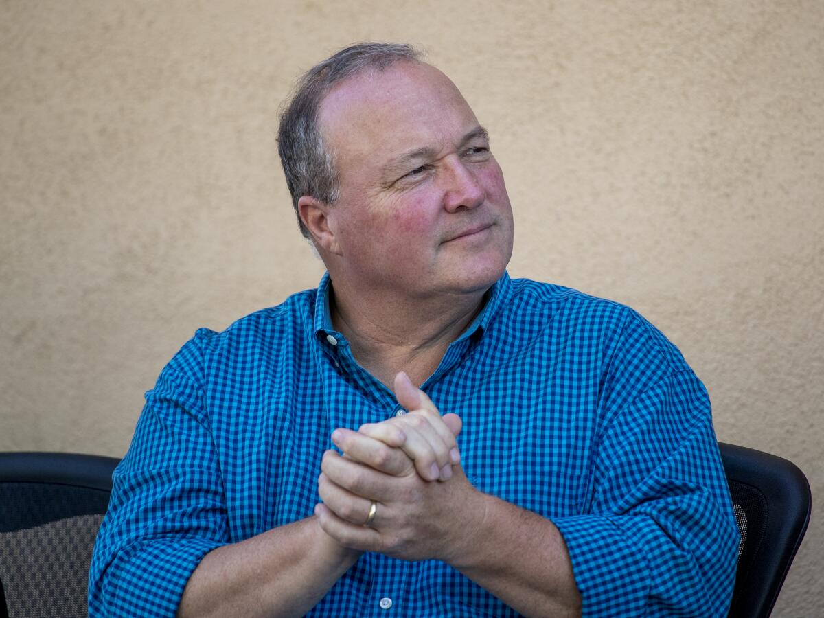 A man in a blue checkered shirt clasps his hands together in front of a beige wall 