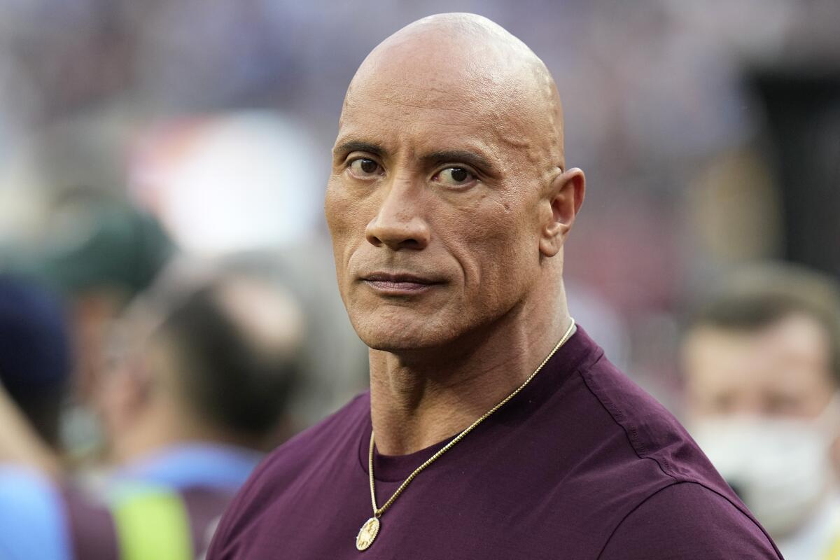 Dwayne "The Rock" Johnson is standing on a football field while wearing a maroon shirt, gold chain with a serious face