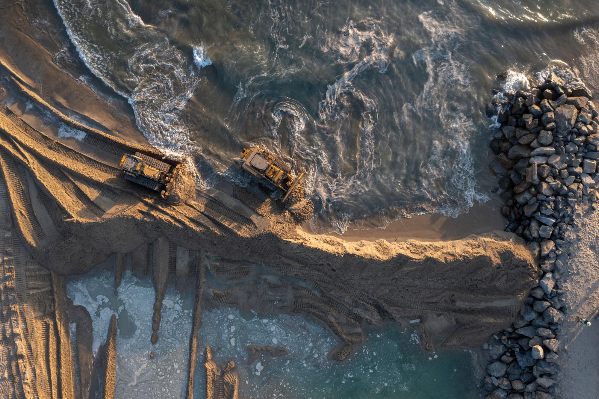An aerial picture of sand berms and heavy equipment near swirling waves and a rocky barrier