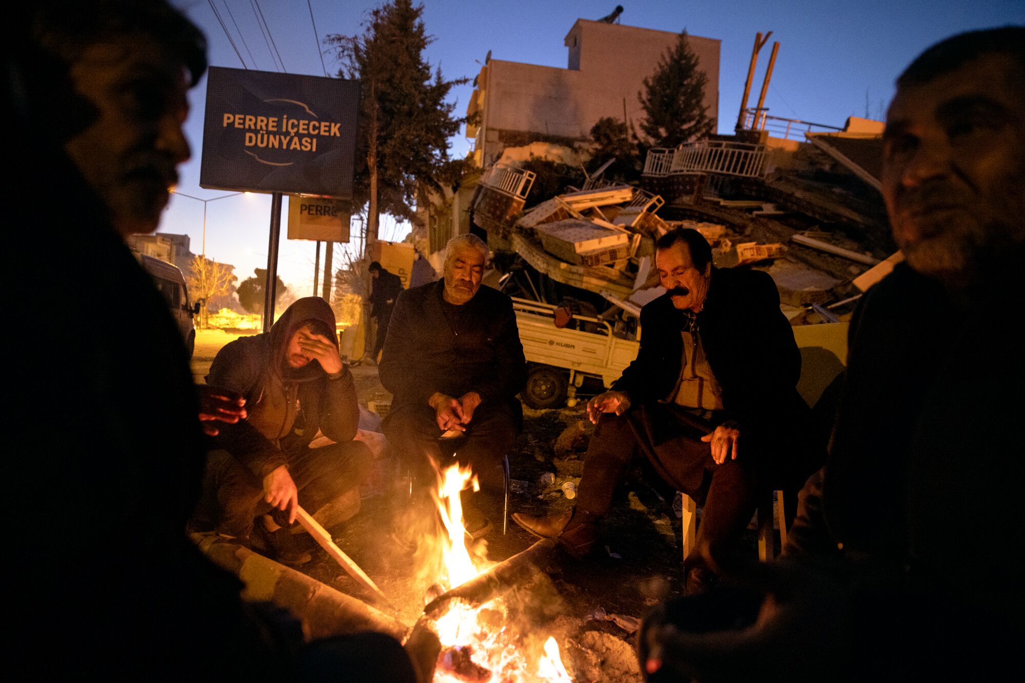 Five men, looking tired and sad, sit around a campfire at night. A collapsed concrete building is behind them.