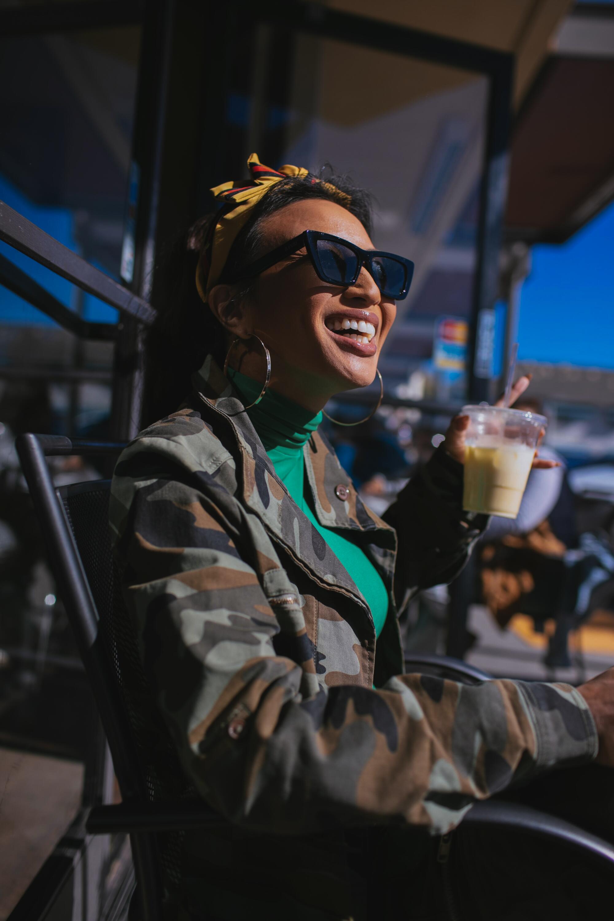 A woman in sunglasses smiles while holding a drink.