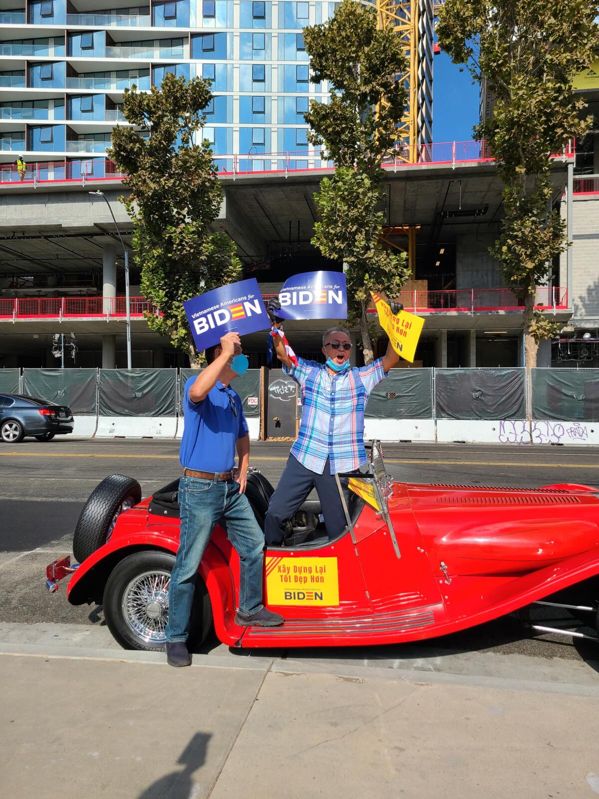 Two people with Biden signs stand next to a small, old-school red car