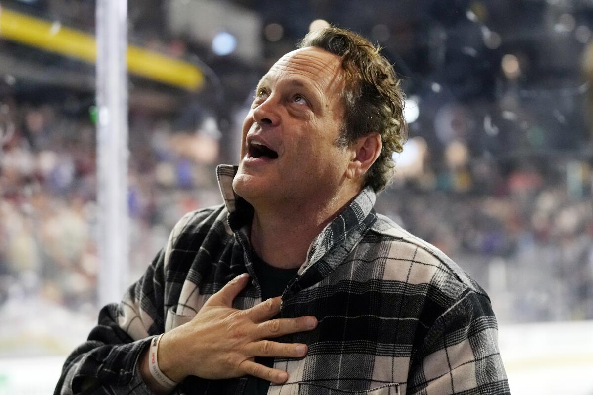 Vince Vaughn, in a plaid button-front shirt, touches his chest as he speaks at a hockey stadium.