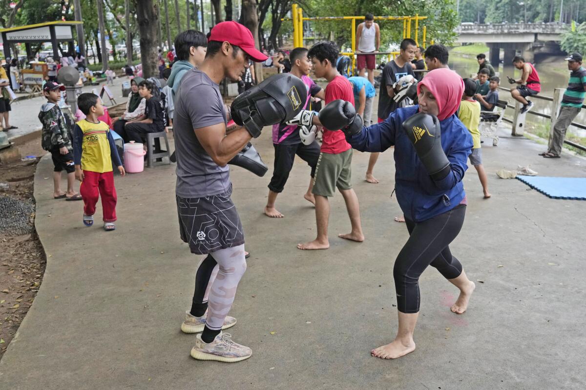 A woman wearing boxing gloves and a bright hijab punches sparring pads held by a trainer at a park in Jakarta, Indonesia.