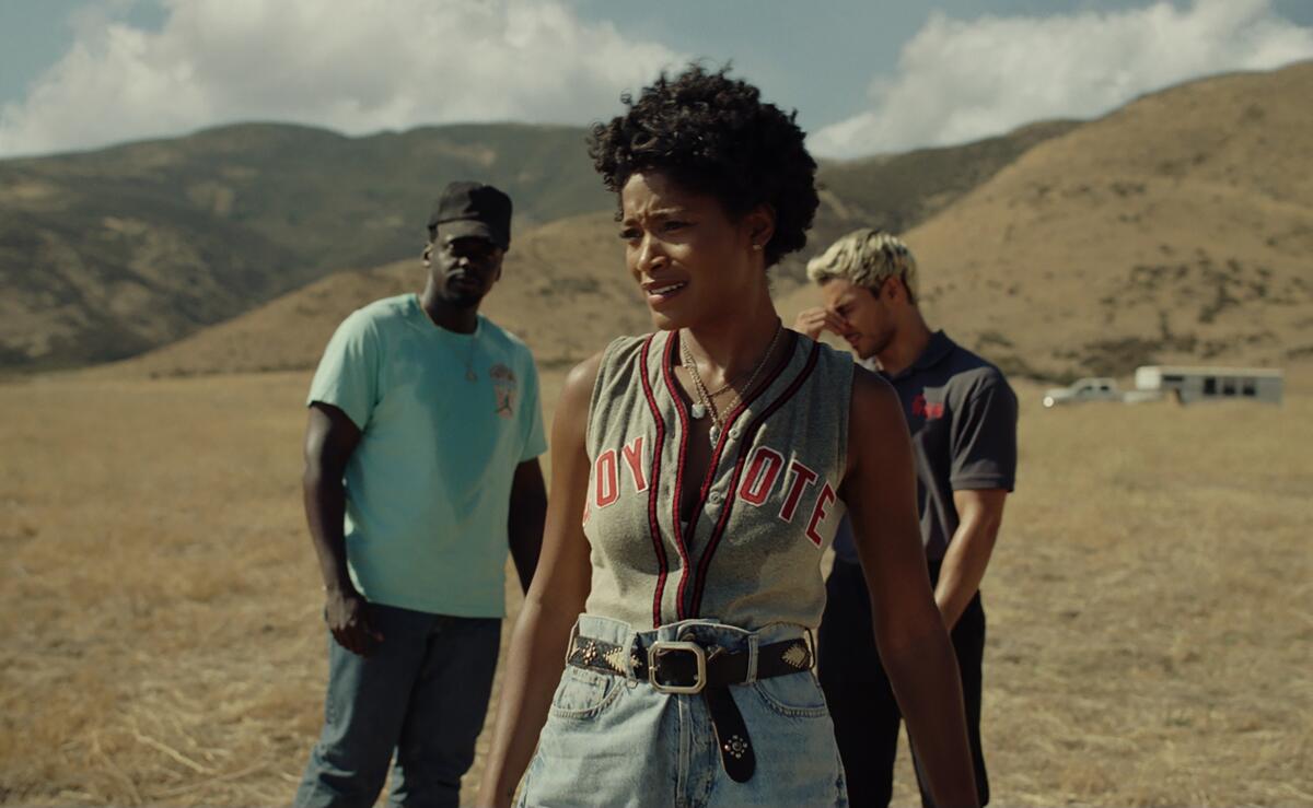 Two men in T-shirts stand behind a woman with short dark hair in an open, empty field with mountains in the background.