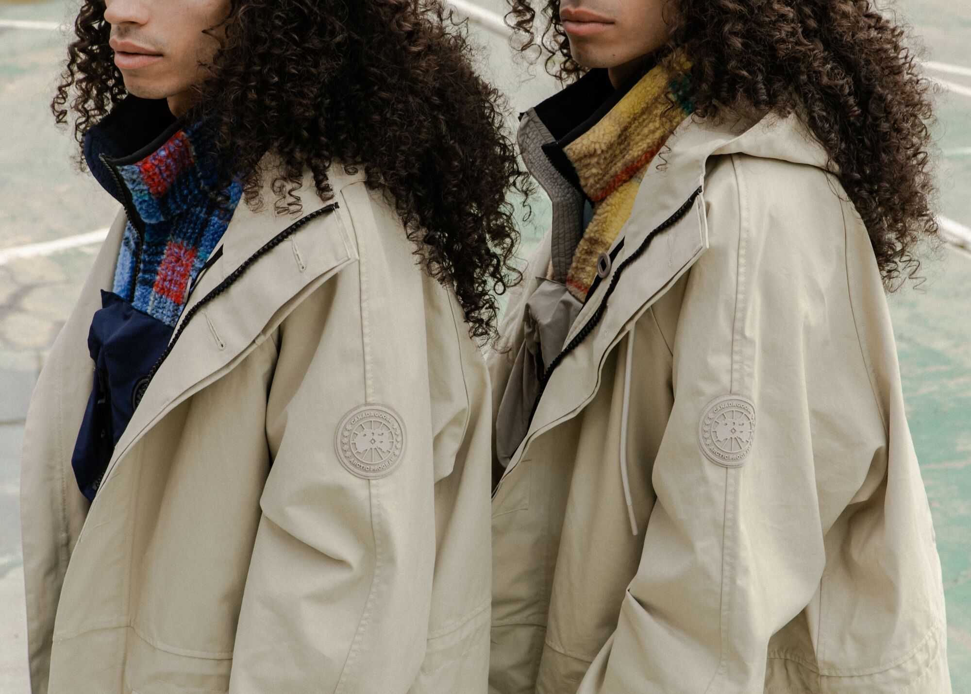 A close-up on the torsos of two models wearing beige parkas with a logo on the upper left shoulder.