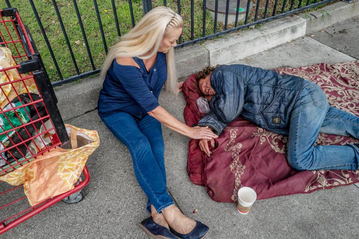 A woman sits next to a man sleeping on blankets on a sidewalk