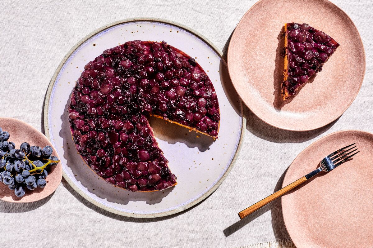 A round fruit-topped cake with a slice of it on an adjacent plate and a bunch of grapes nearby.