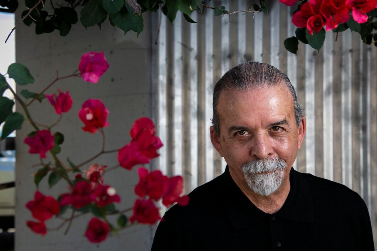 A man in a black shirt and a gray goatee sits before flowers and an aluminum wall panel.