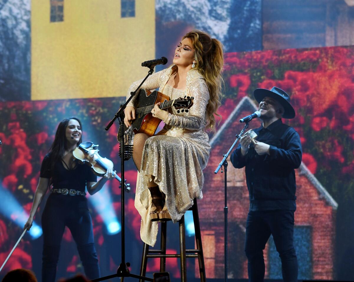 Shania Twain, sitting on a stool, plays the guitar and sings. Behind her are two background singers.