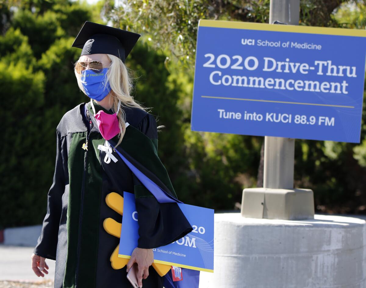 A UC Irvine administrator wearing cap, gown and mask walks past a sign announcing a "drive-through commencement"