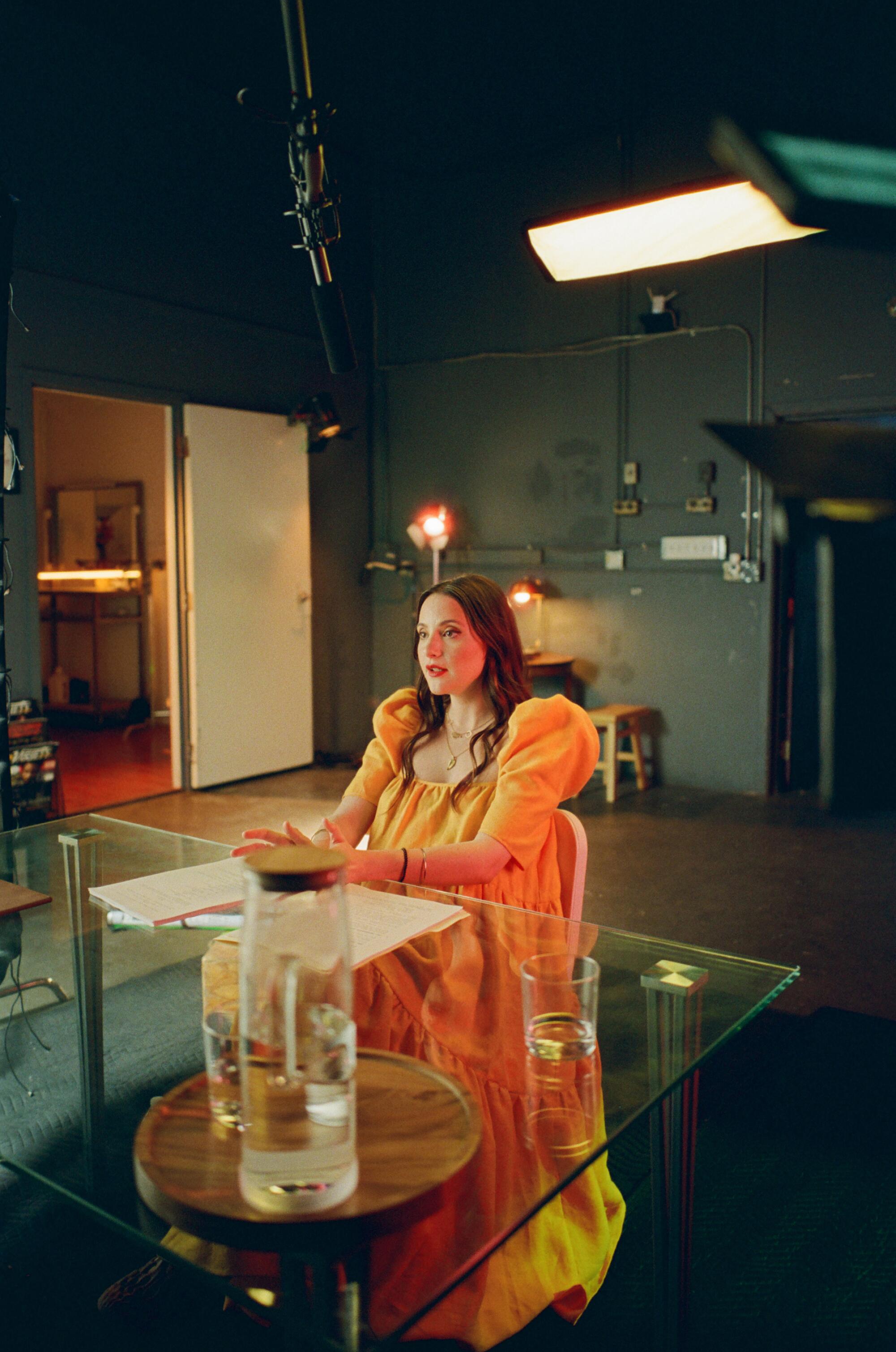 Alexa Nikolas, in an orange dress with puffy sleeves, sits at a glass table on set.