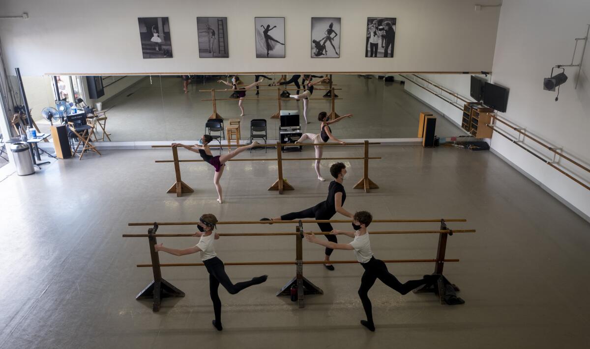Five young dancers wearing masks rehearse at a barre in a dance class. 