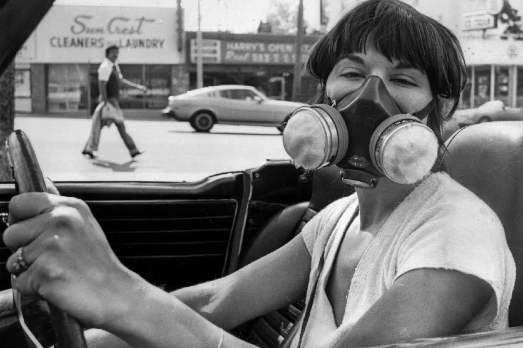 A woman sits at the wheel of a convertible wearing a gas mask