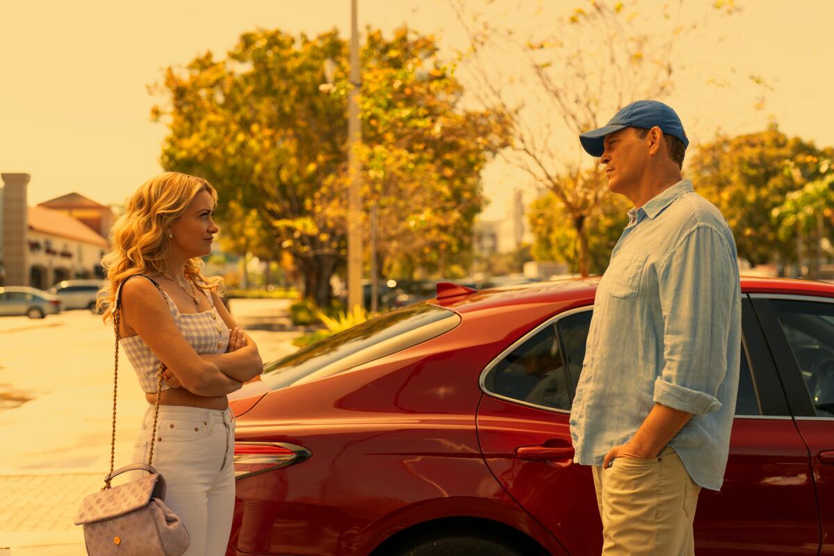 A woman stands next to a red car with her arms crossed looking at a man in a baseball cap.