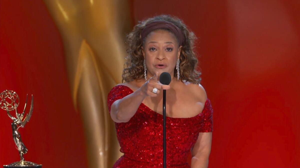 Debbie Allen, in a sparkly red dress, speaks into a microphone and points to her audience
