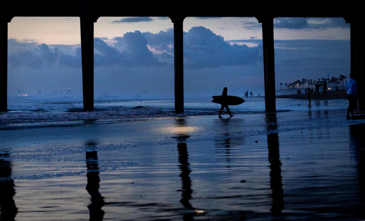 A surfer is silhouetted at dusk beneath a pier.