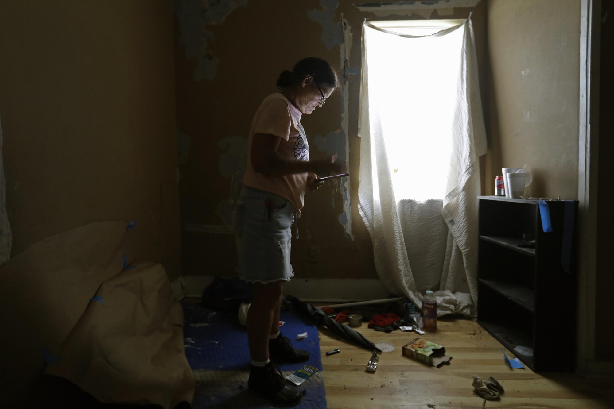 A woman stands in a room with sheer white window coverings and debris littering the floor inside a home