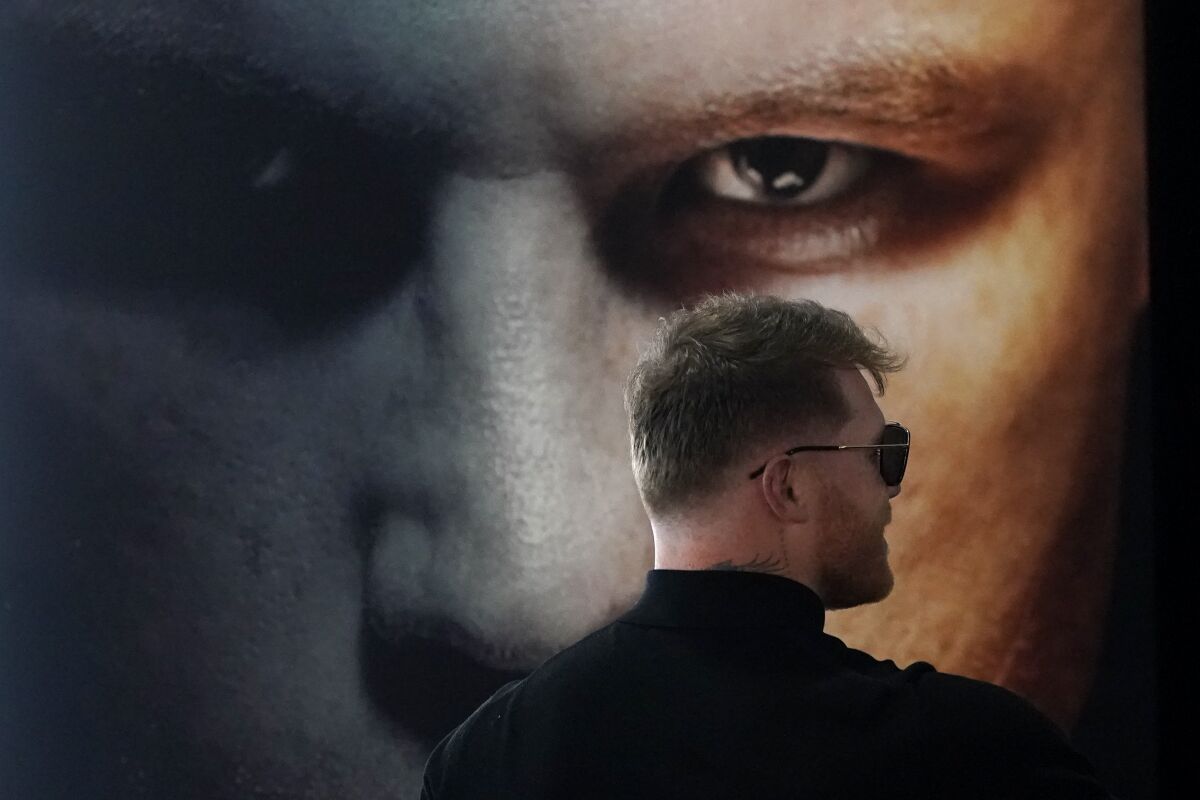 Boxer Canelo Alvarez wears sunglasses and stands facing a poster promoter his fight with Dmitry Bivol.