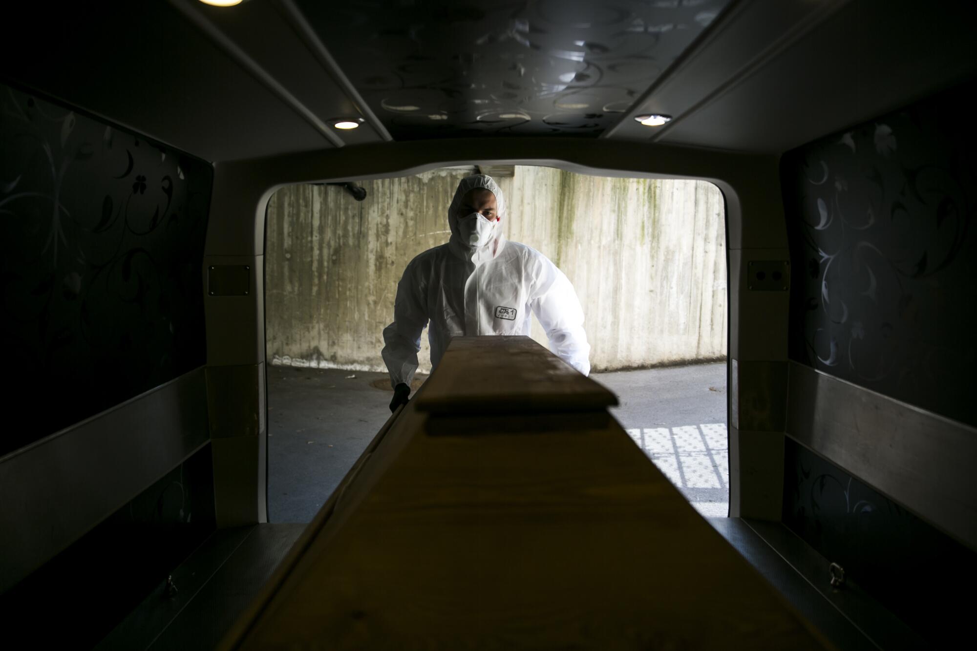 An undertaker in full protective clothing pushes a coffin into a mortuary van.