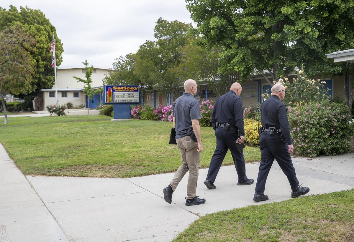 Two police officers and a man walk at right toward an entrance of a building with a sign at left that reads "Saticoy"