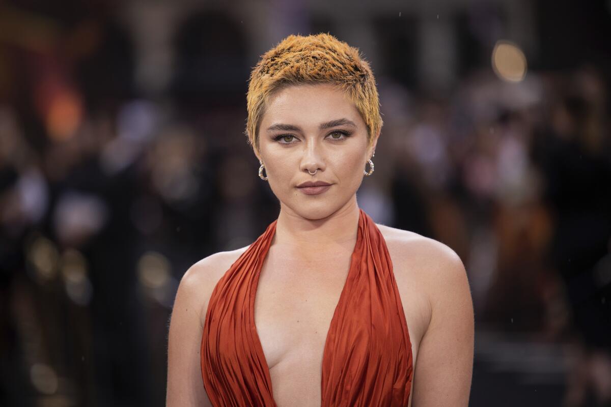 Florence Pugh poses with orangy buzzcut hair in a rust dress with a plunging neckline.