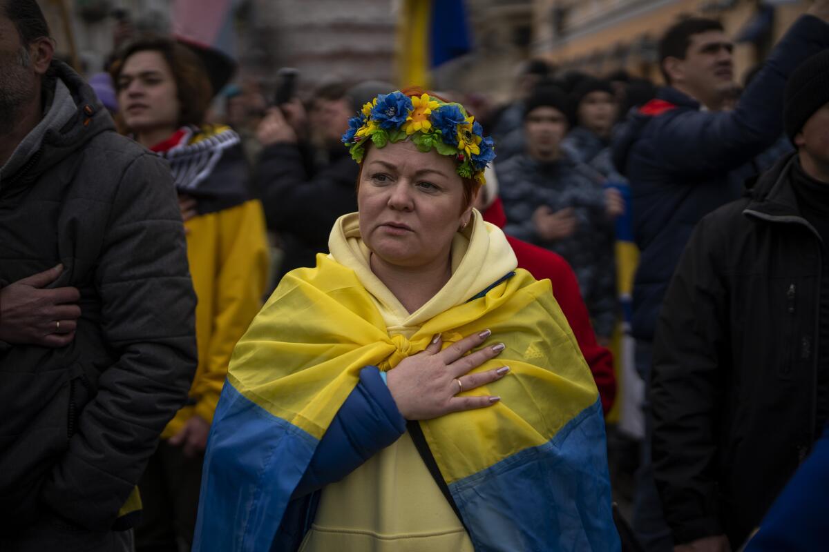 A woman among a crowd sings with hand over heart, flowers on head, a Ukrainian flag around shoulders