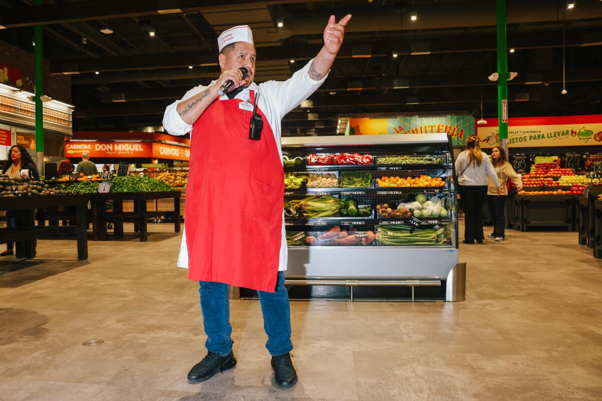 A man in a red apron holding a microphone, standing in the middle of the produce section of a grocery store.