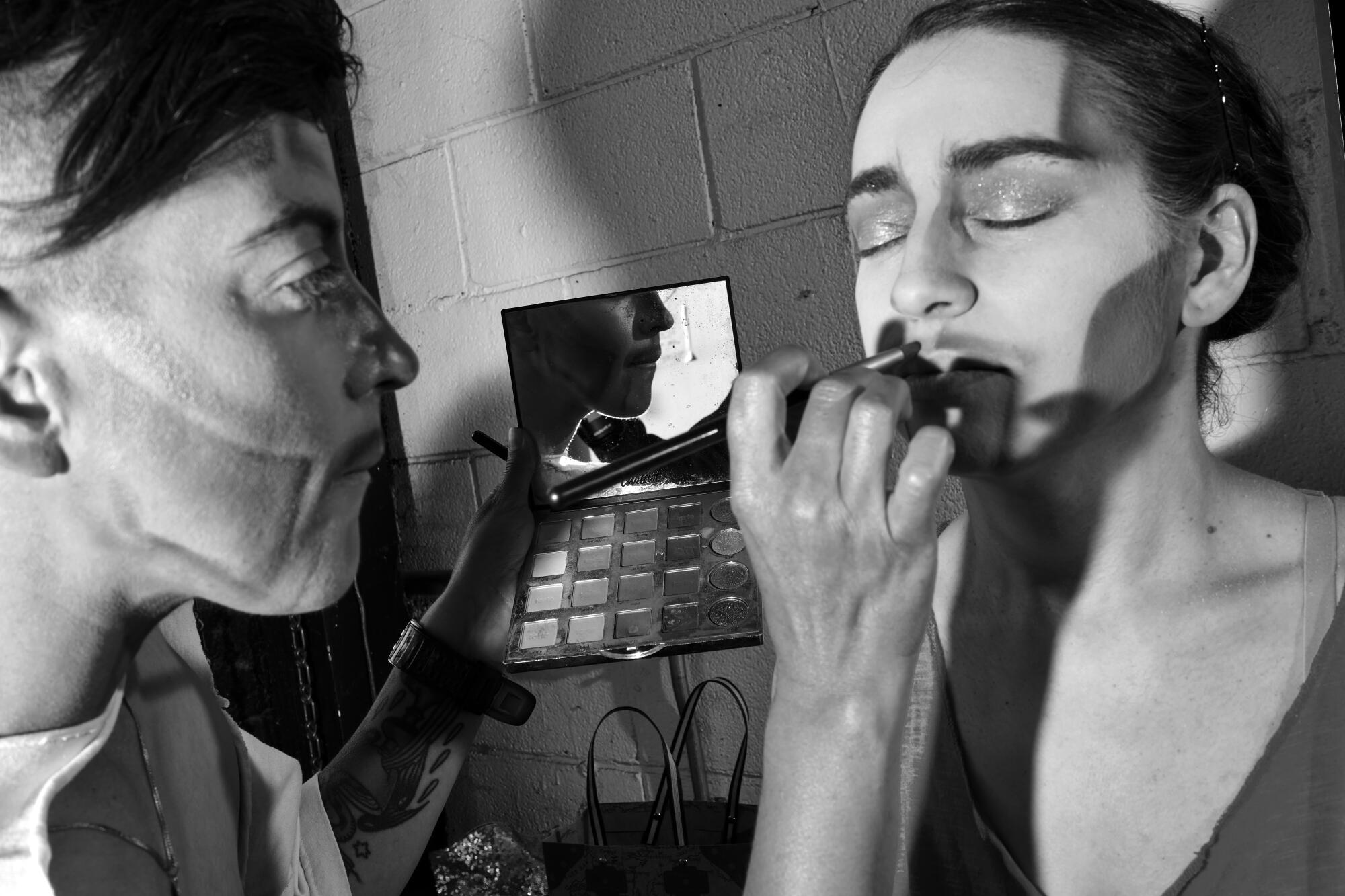 A person with a made-up face applies makeup to another person in a black-and-white photo