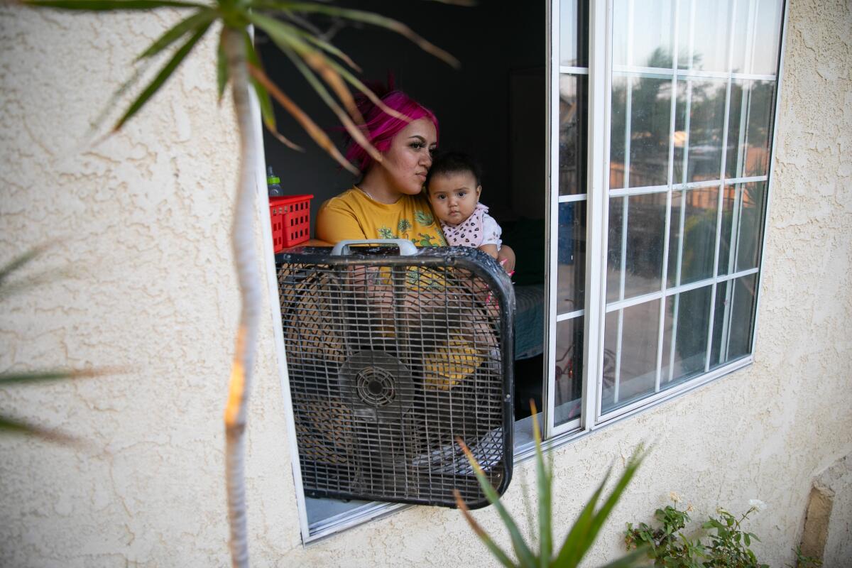 A woman stands in front of a fan in an open window, holding her baby daughter, to try to cool off on a hot day.