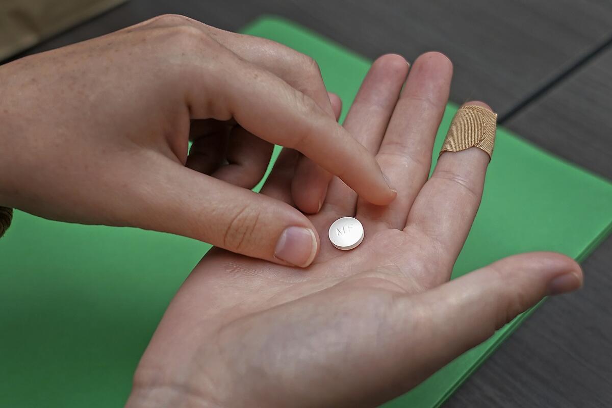 A closeup of a person's left hand poised to pick up a round white pill from the palm of their open right hand.