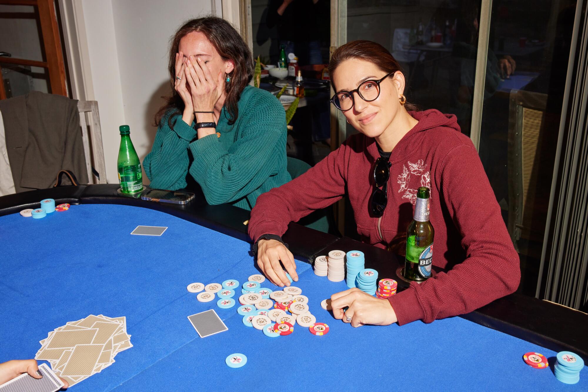 Two women seated at a poker table, one covering her face, the other raking in the chips
