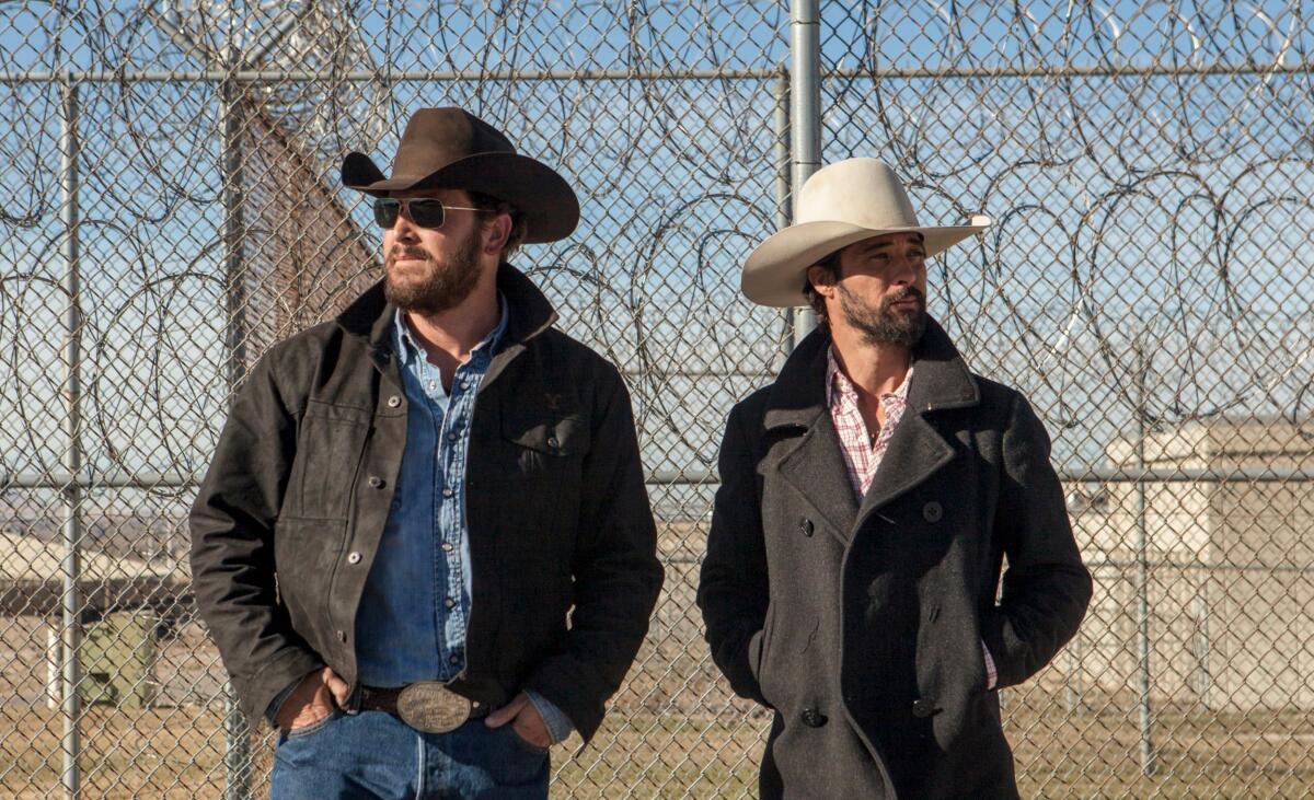 Two men in cowboy hats stand in front of a chain-link and razor-wire fence.
