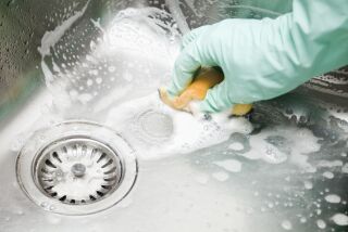 Gloved hands scrub a large stainless-steel kitchen sink with a scouring pad, soap bubbles.