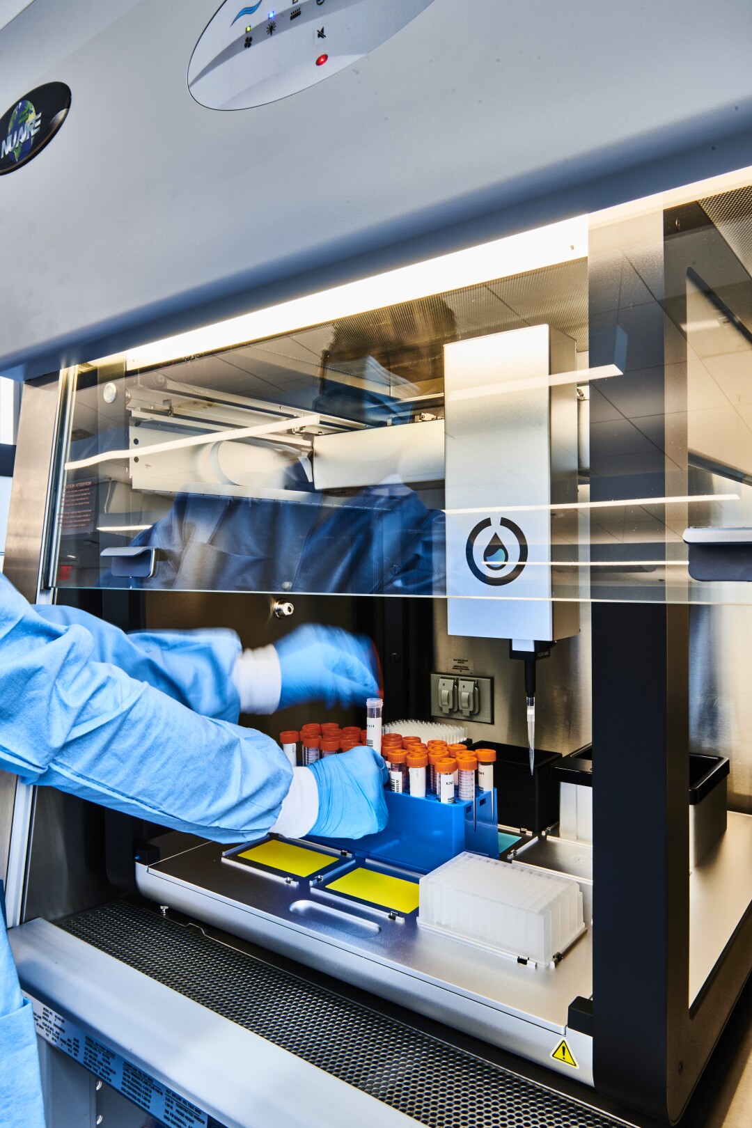 A pair of hands holds one of a group of test tubes with red caps placed inside a rectangular machine with clear sides