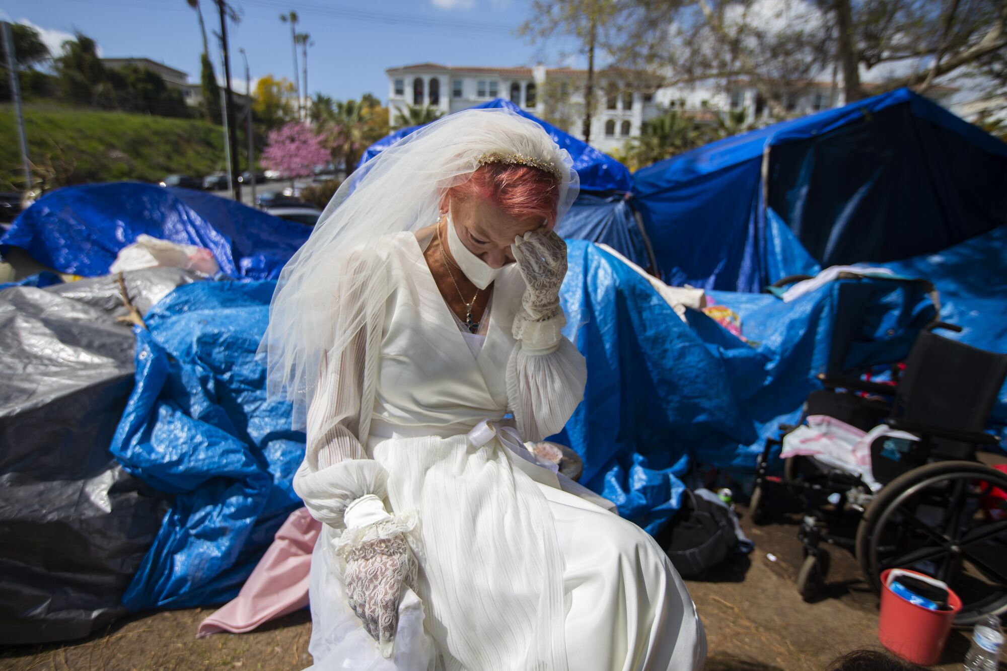 A woman in a wedding dress sits among tents in a park.