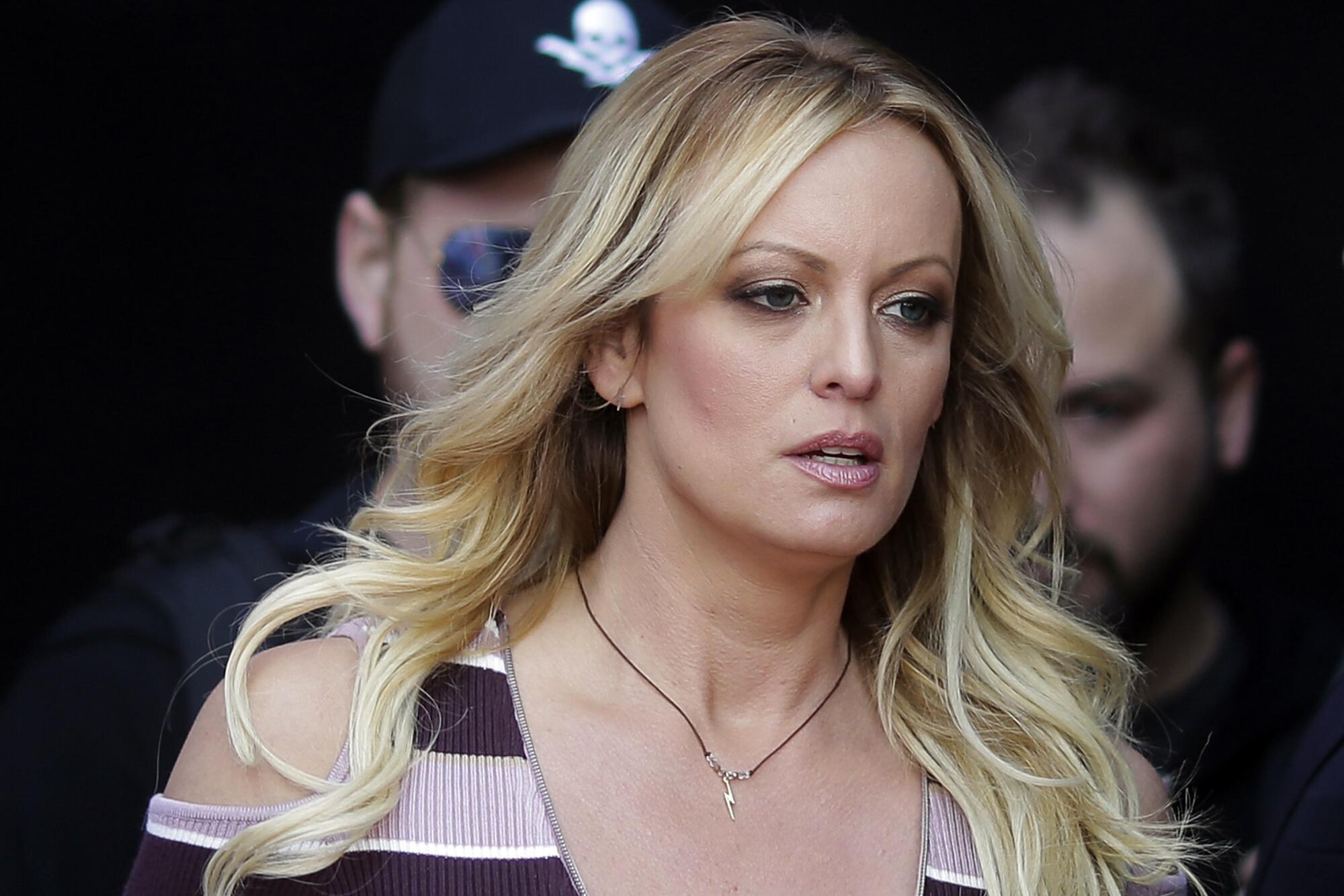 Stormy Daniels pictured from the shoulders up as she walks past two men in the background.
