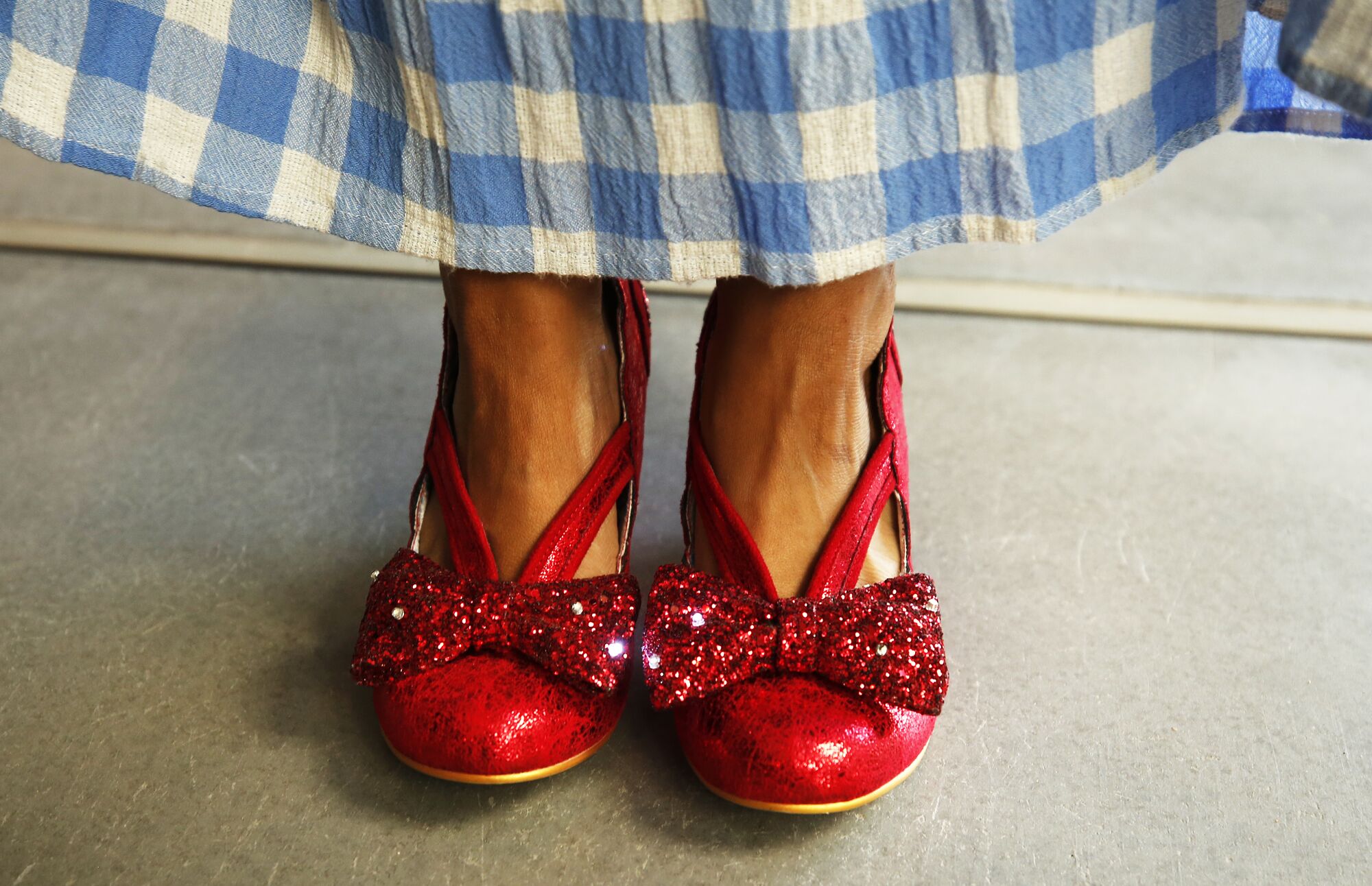 A woman's feet in her own "ruby slippers" below the hem of her blue-and-white gingham skirt.