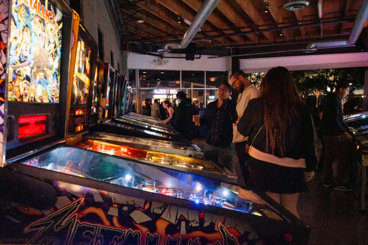 A row of pinball machines in a dark room with several people standing in front of them.