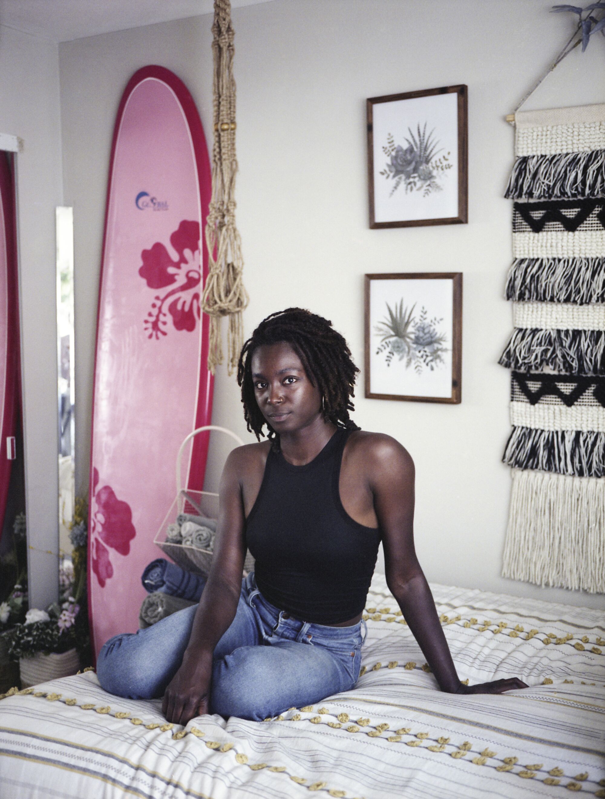 A surfer sits on her bed, a pink surfboard behind her.