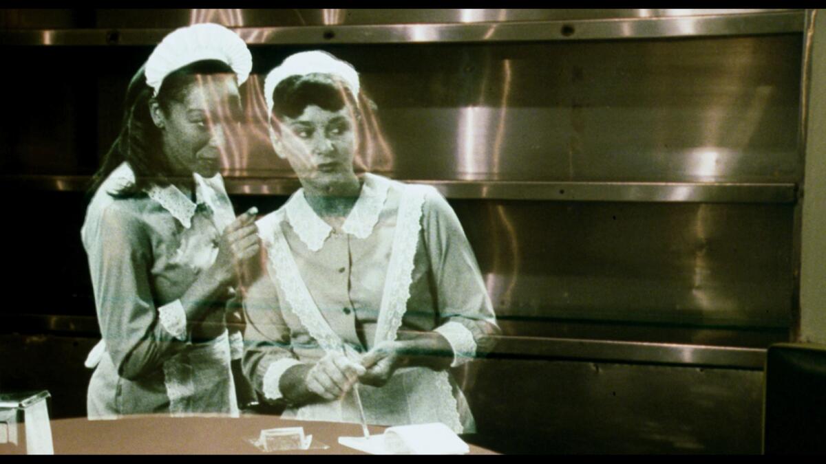 A still shows ghostly black-and-white images of two women in housekeeper costumes whispering to each other in a kitchen.